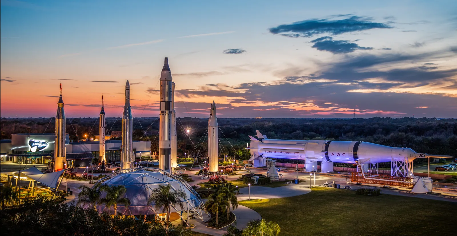 Rocket Garden at dusk seen from above, with lit rockets, a Saturn IB lying on its side and the visitor complex buildings
