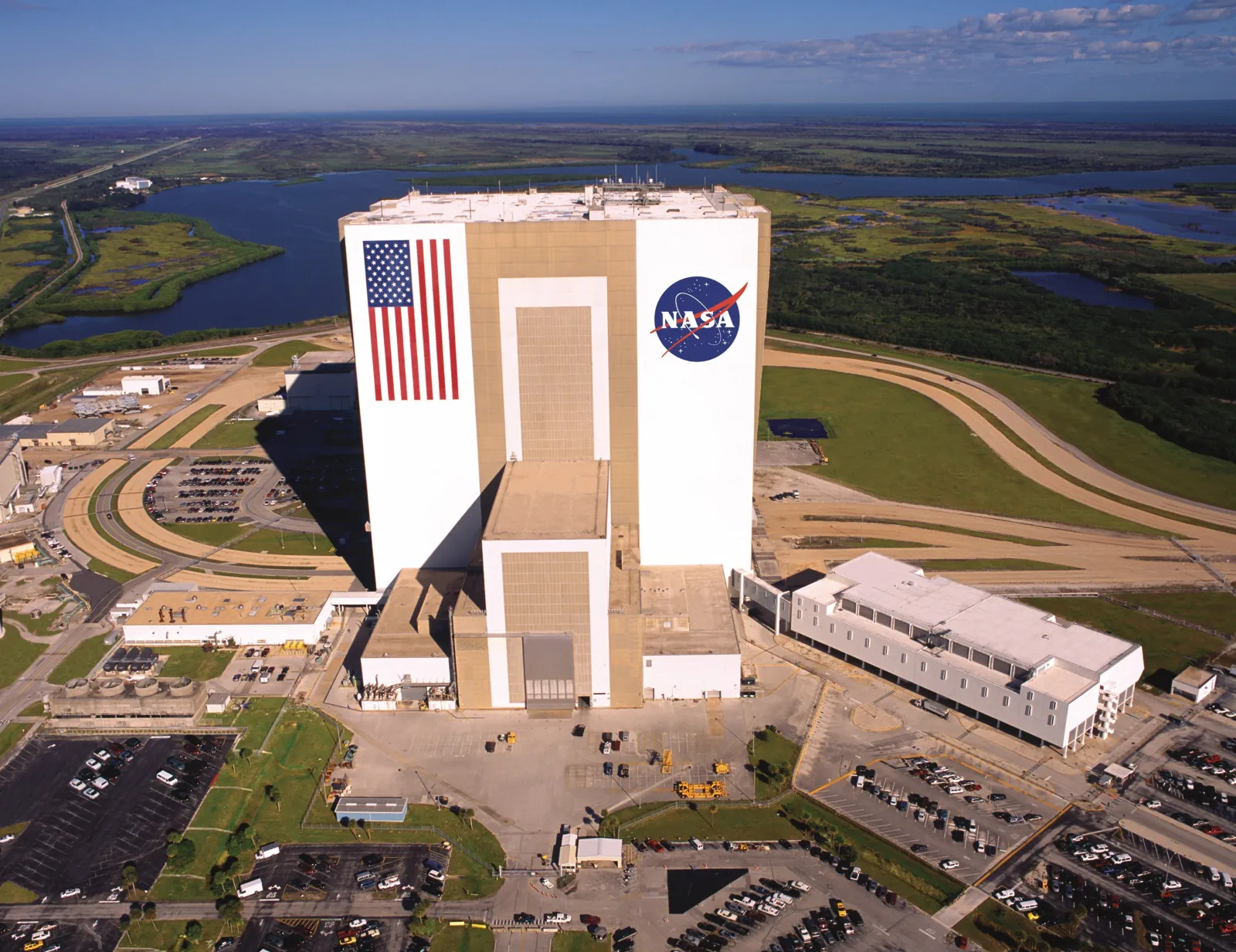 Aerial view of the Vehicle Assembly Building with its American flag and NASA logo, surrounded by wetlands and lagoon