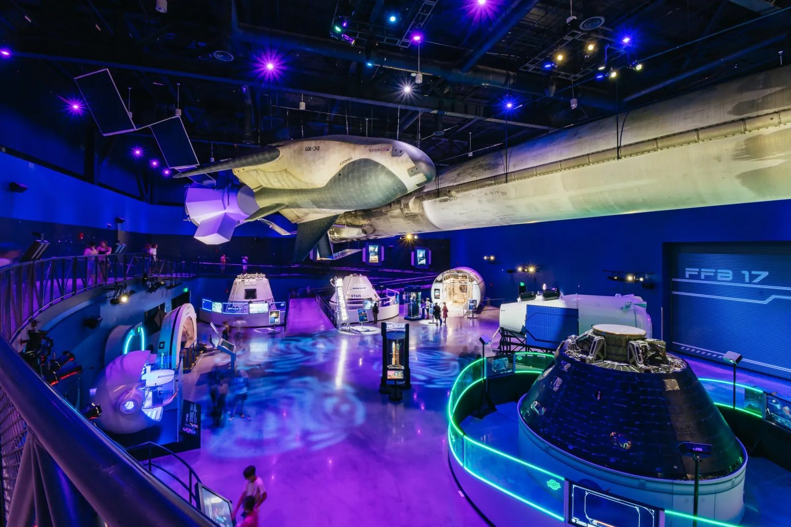 Gateway exhibit hall lit in purple and blue, with a Dream Chaser spaceplane overhead and crew capsules on the floor
