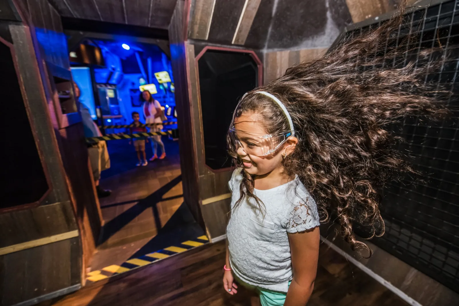 Girl in safety goggles with her hair blown sideways inside the hurricane wind exhibit