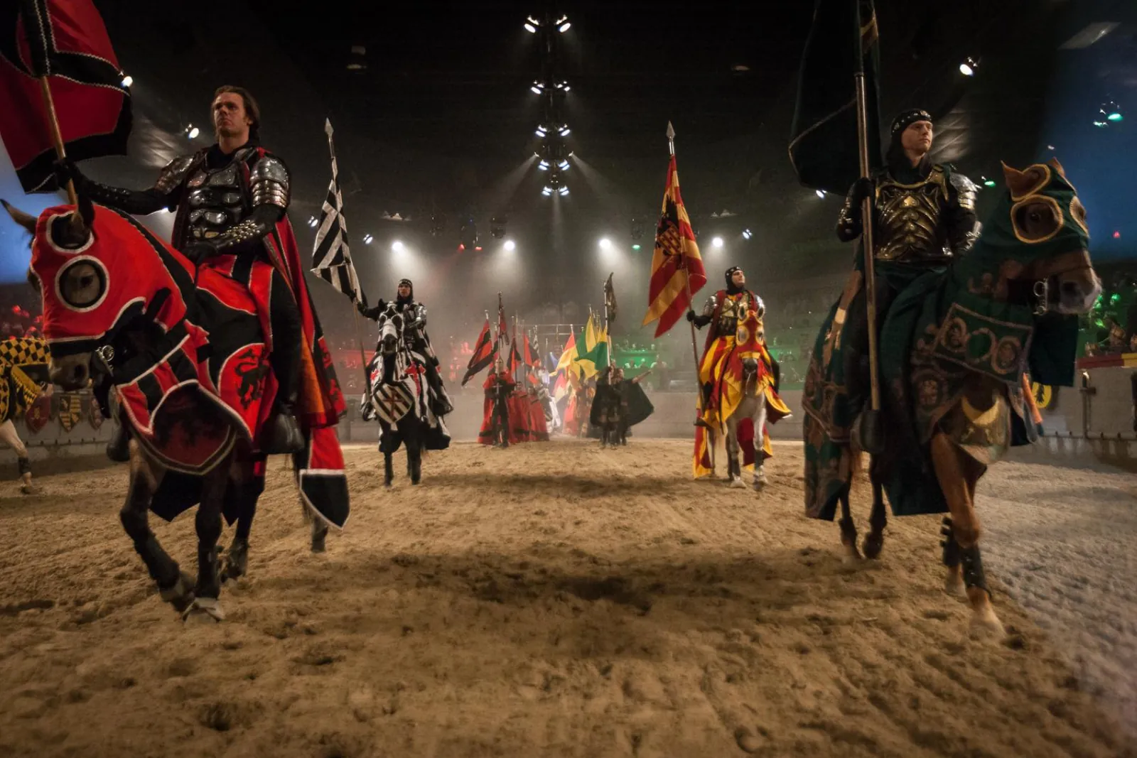Knights on caparisoned horses carrying banners across the sand arena under stage lights