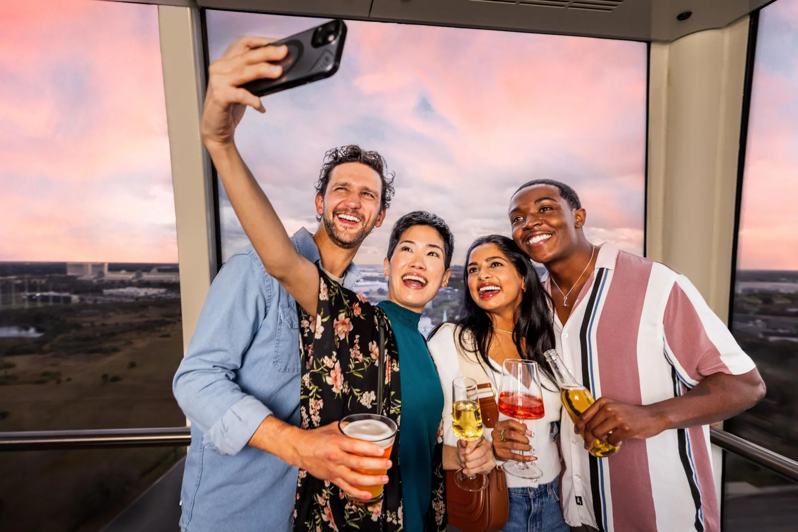 Four friends taking a selfie inside a glass capsule with a pink sunset sky behind them