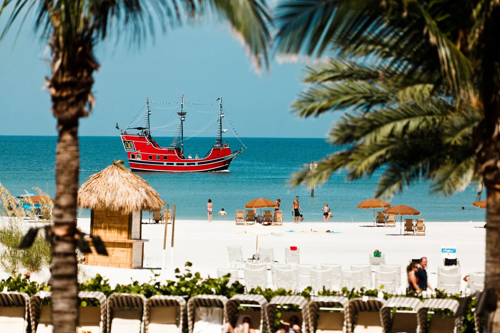 Red pirate ship sailing past Clearwater Beach, framed by palm trees and a thatched hut