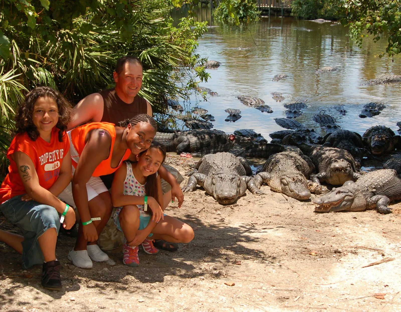 Smiling family crouched on a sandy bank beside a group of basking alligators