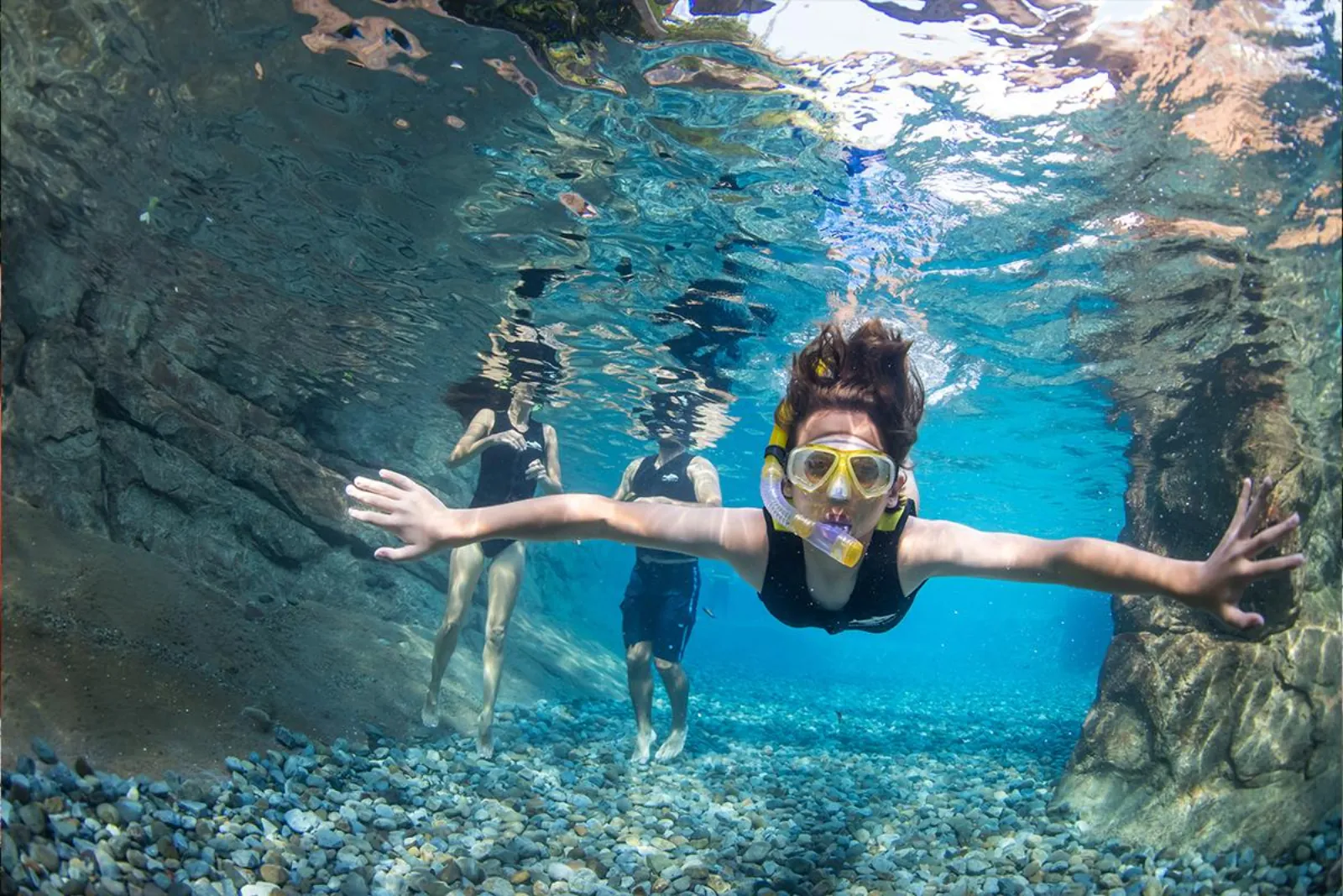 Child snorkelling underwater in a clear rocky channel with two adults wading behind
