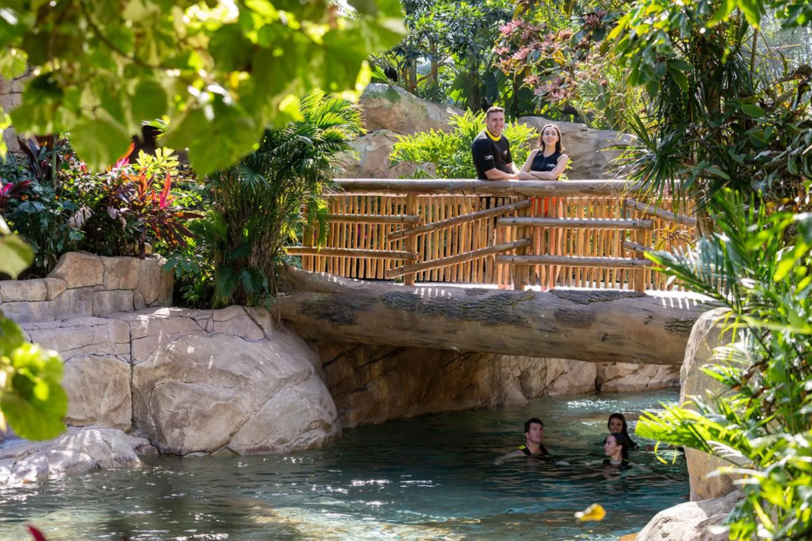 Couple on a wooden bridge above swimmers in a shaded, jungle-lined river