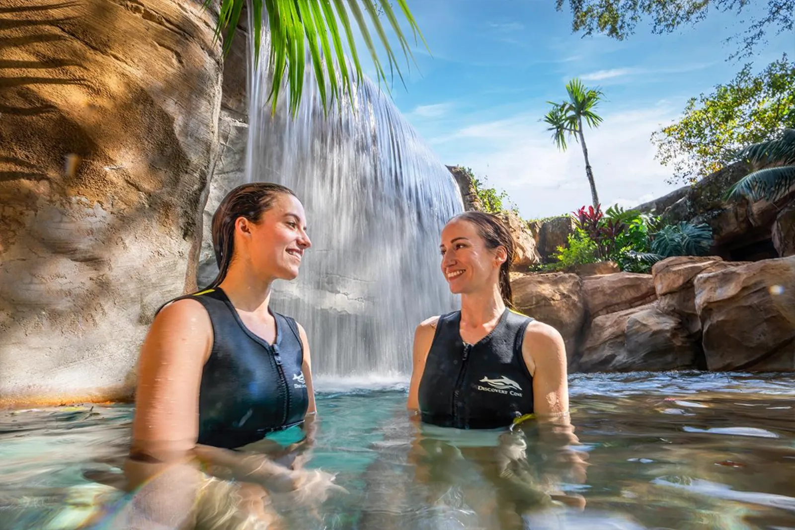 Two women in Discovery Cove wetsuits standing in the water in front of a waterfall