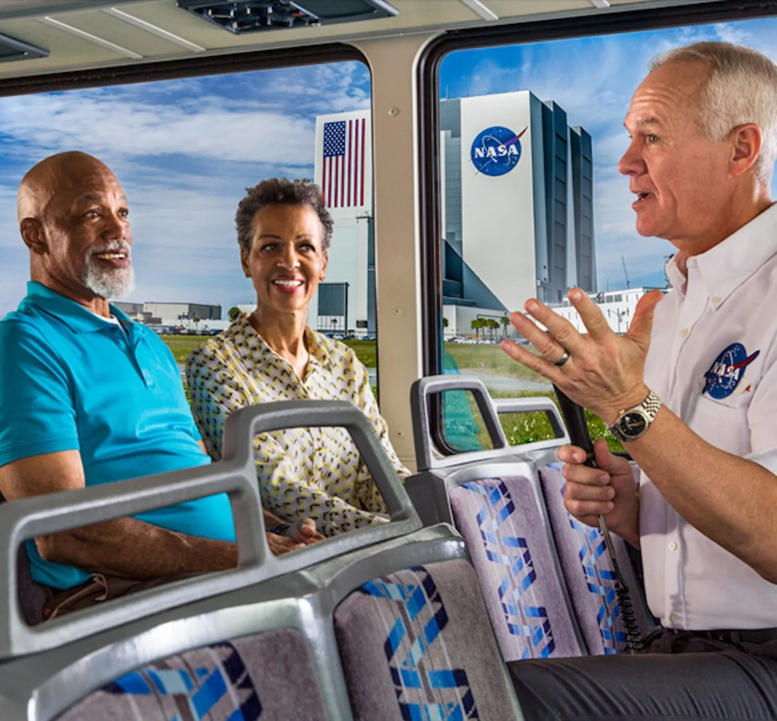 Guide in a NASA shirt talking to passengers on a tour bus, with the Vehicle Assembly Building visible through the window