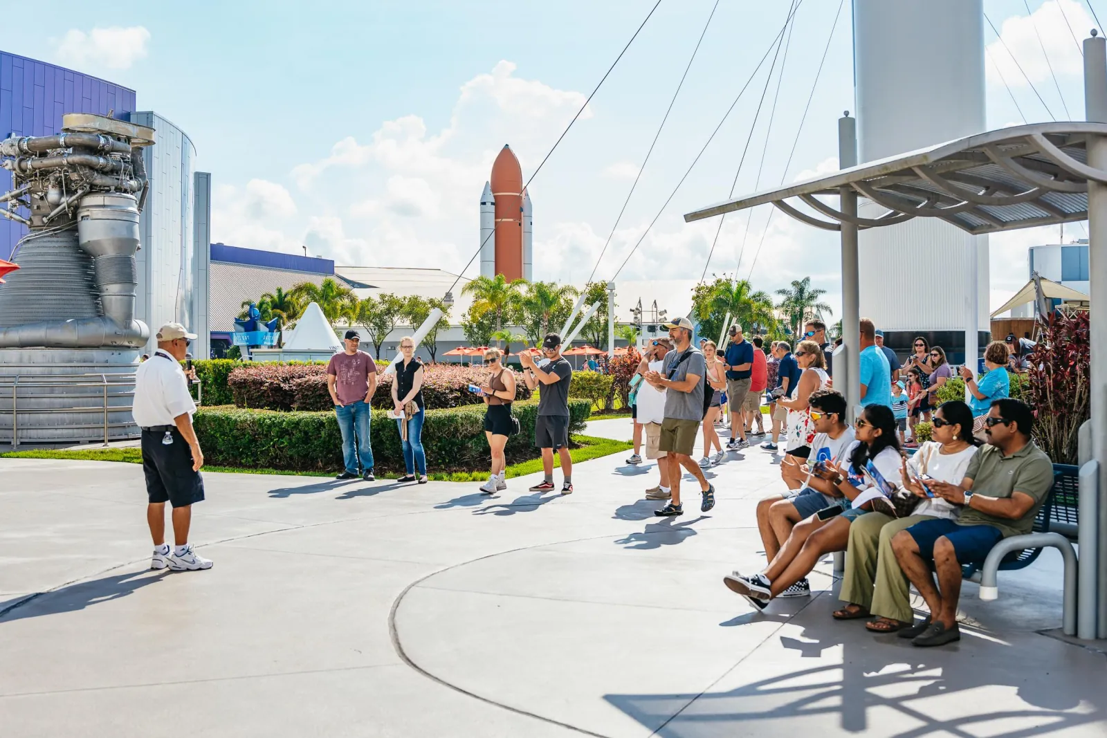 Staff member speaking to a group of visitors in the Rocket Garden, with the shuttle external tank and boosters display behind