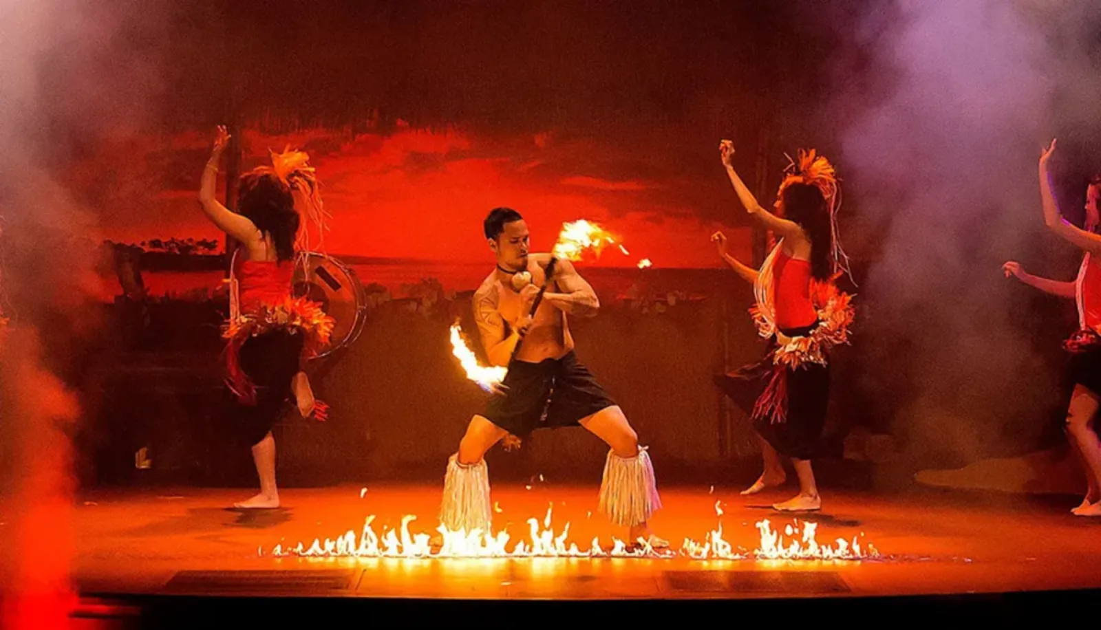 Fire-knife dancer crouching behind a line of flames on stage between dancers in feathered costumes