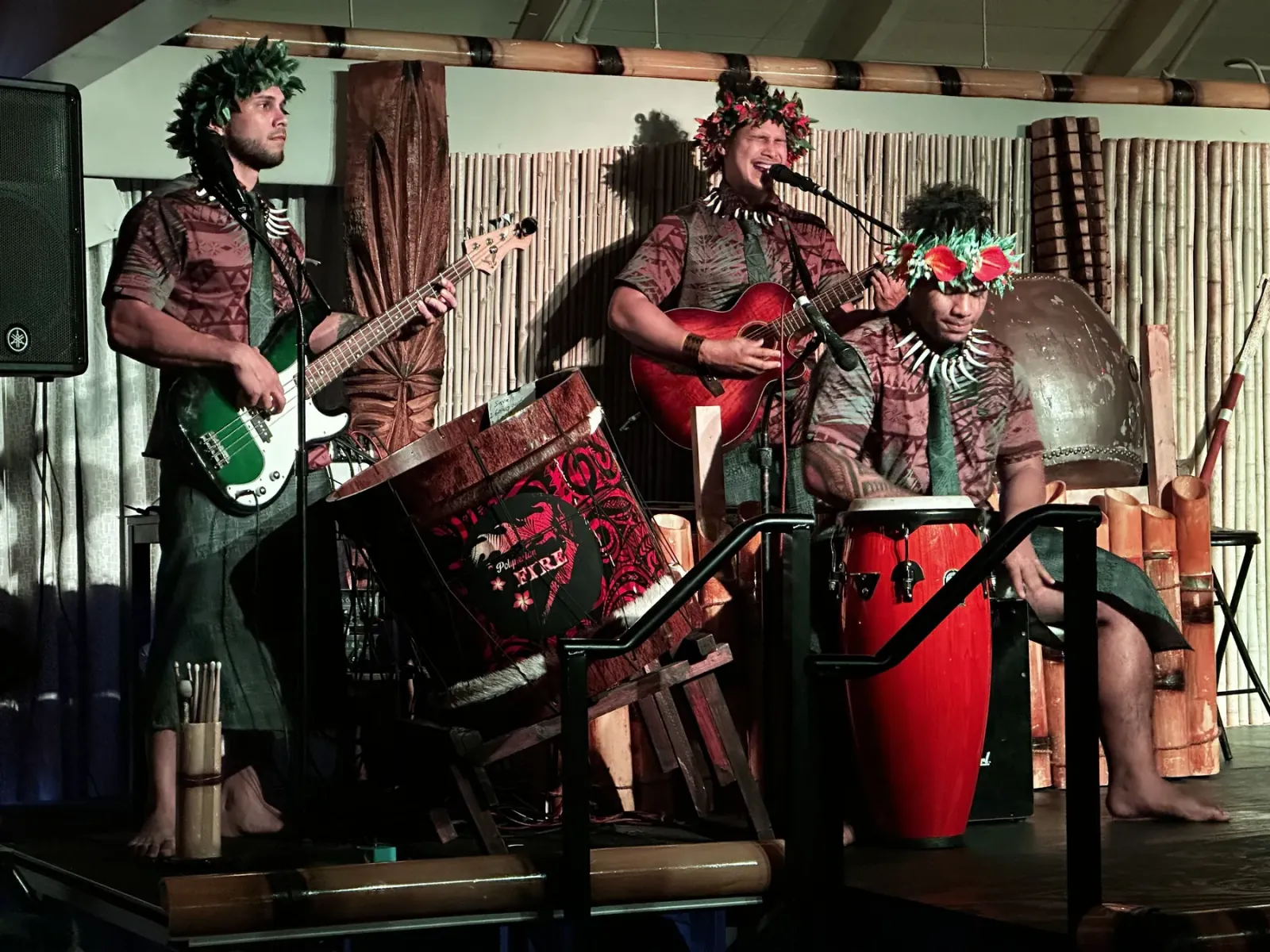Three musicians in floral head wreaths playing bass, guitar and drums on the luau stage