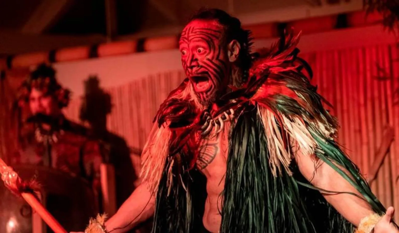 Performer in face paint and a feathered cloak performing a warrior chant under red light