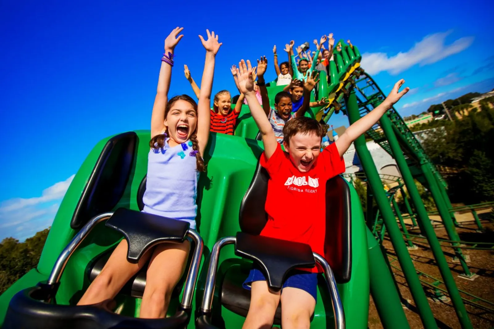 Children with arms raised riding a green LEGOLAND roller coaster under a blue sky
