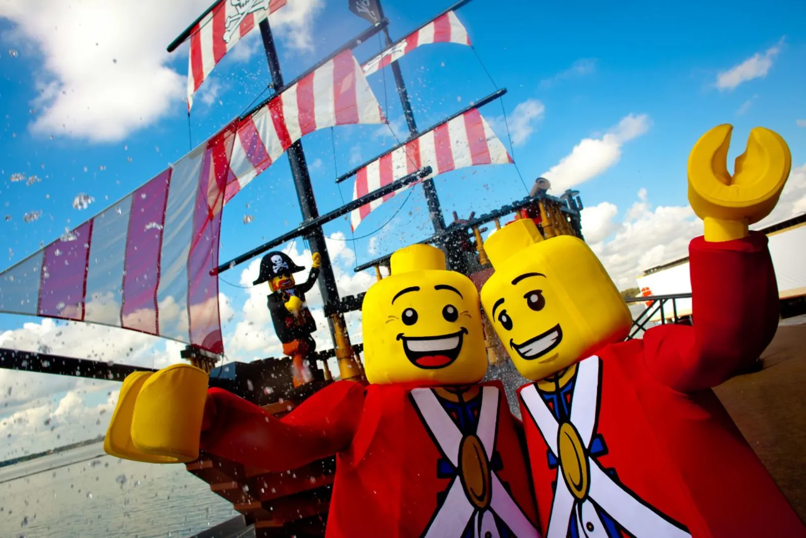Two costumed LEGO minifigure characters waving in front of a pirate ship
