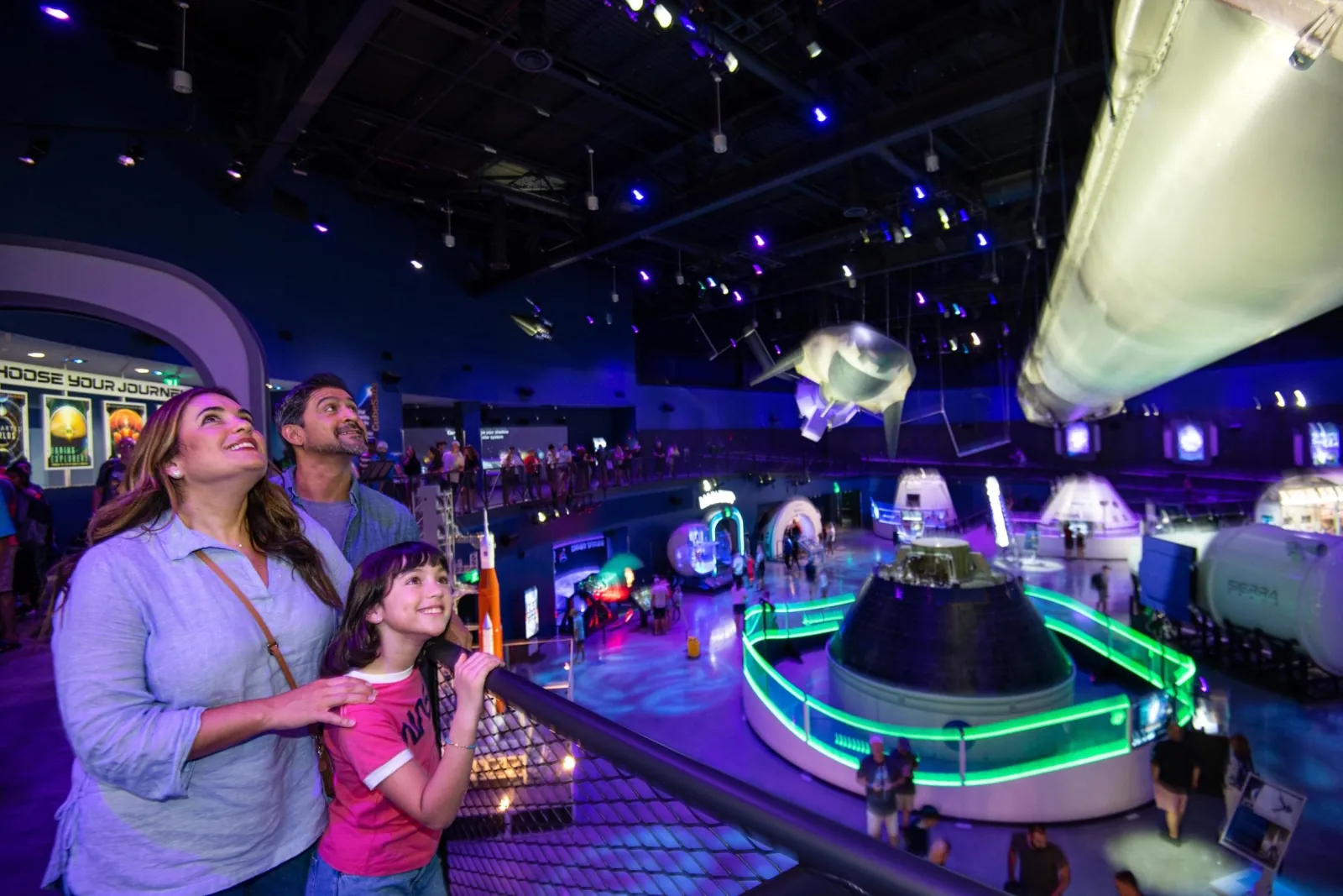 Family at a balcony railing looking over the Gateway exhibit hall, with crew capsules below and a spacecraft hanging overhead