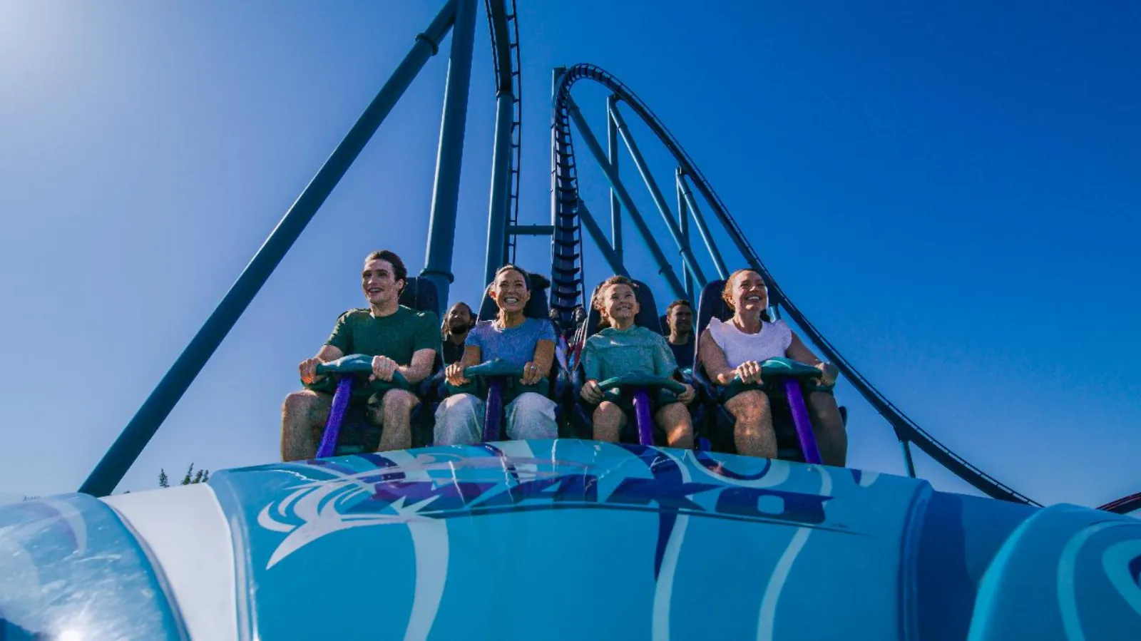 Riders smiling in the front row of the Mako roller coaster, with its steep blue track against the sky