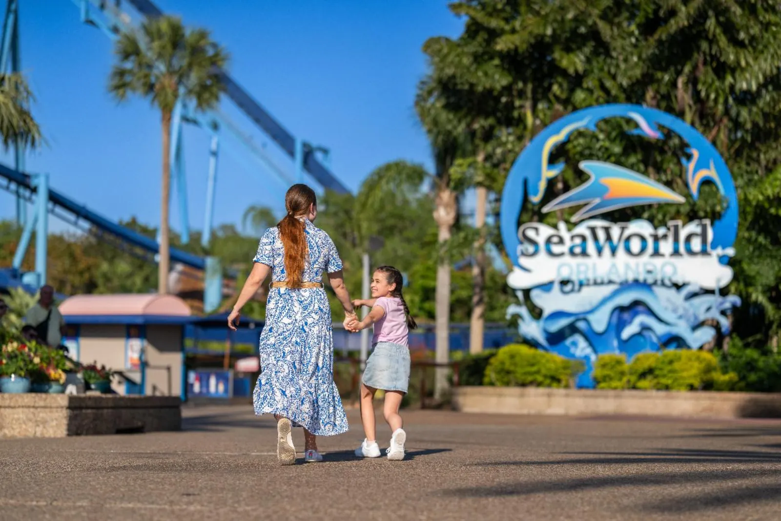 Mother and daughter walking hand in hand toward the SeaWorld Orlando entrance sign, with blue coaster track behind