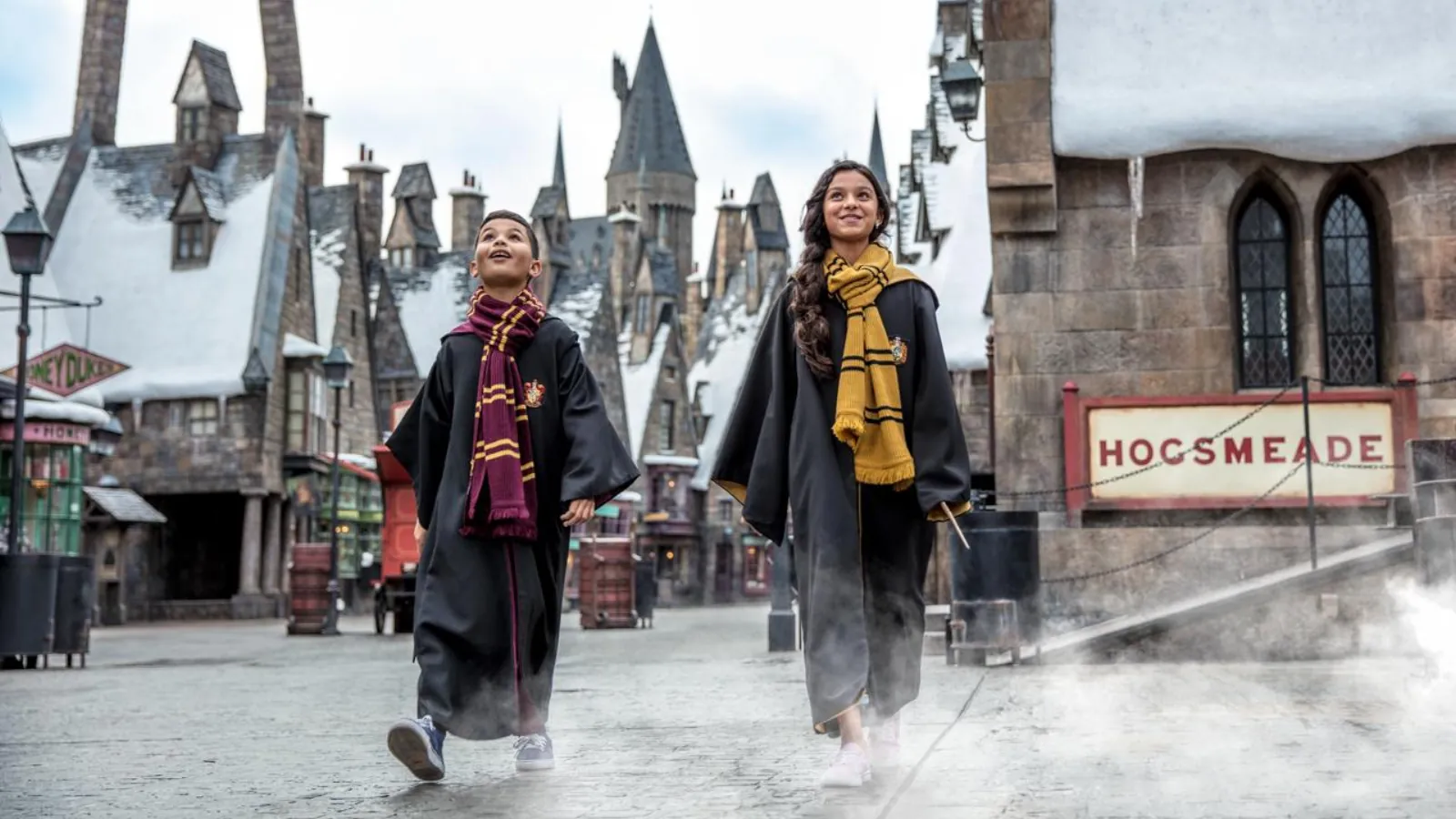 Two children in Hogwarts robes and house scarves walking through snow-roofed Hogsmeade